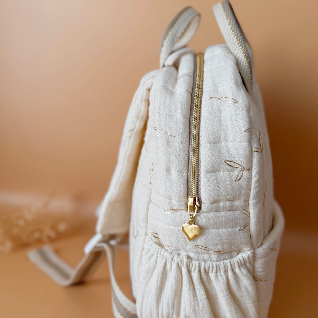 Voici notre tout nouveau sac à dos maternelle, pensé et confectionné en France pour accompagner vos tout-petits dans leurs premières journées d'école. Ce modèle délicat est réalisé en gaze de coton matelassée écrue, ornée de petites feuilles dorées qui lui donnent une allure douce et poétique. La grande particularité de ce sac ? Il est personnalisé avec le prénom de votre enfant, brodé à la main en fil doré sur la poche avant. Un détail unique, raffiné, et tellement précieux pour marquer les affaires de vot