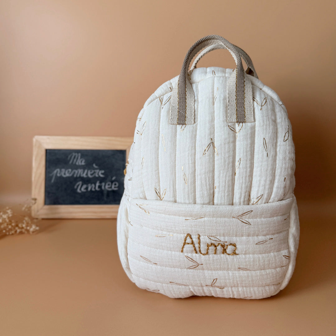 Voici notre tout nouveau sac à dos maternelle, pensé et confectionné en France pour accompagner vos tout-petits dans leurs premières journées d'école. Ce modèle délicat est réalisé en gaze de coton matelassée écrue, ornée de petites feuilles dorées qui lui donnent une allure douce et poétique. La grande particularité de ce sac ? Il est personnalisé avec le prénom de votre enfant, brodé à la main en fil doré sur la poche avant. Un détail unique, raffiné, et tellement précieux pour marquer les affaires de vot