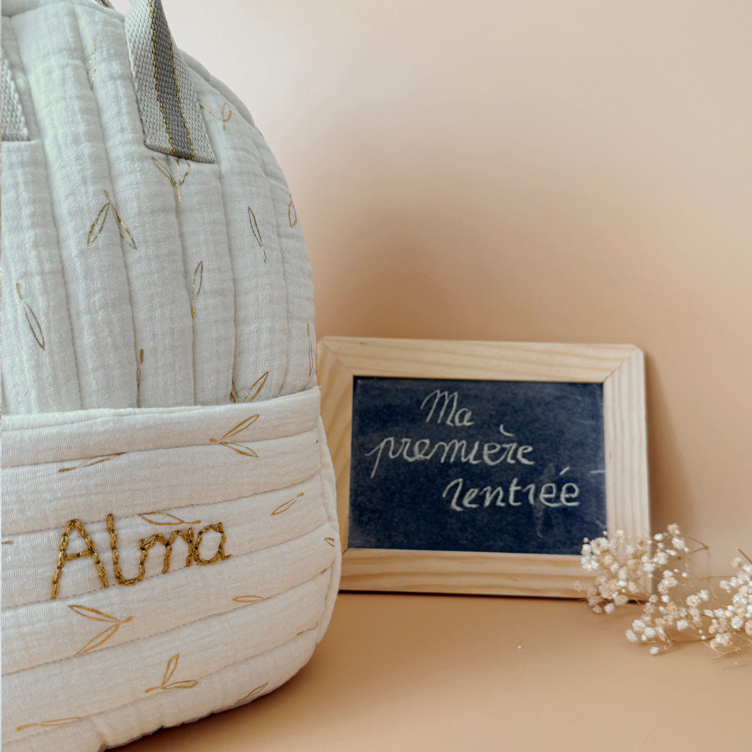 Voici notre tout nouveau sac à dos maternelle, pensé et confectionné en France pour accompagner vos tout-petits dans leurs premières journées d'école. Ce modèle délicat est réalisé en gaze de coton matelassée écrue, ornée de petites feuilles dorées qui lui donnent une allure douce et poétique. La grande particularité de ce sac ? Il est personnalisé avec le prénom de votre enfant, brodé à la main en fil doré sur la poche avant. Un détail unique, raffiné, et tellement précieux pour marquer les affaires de vot