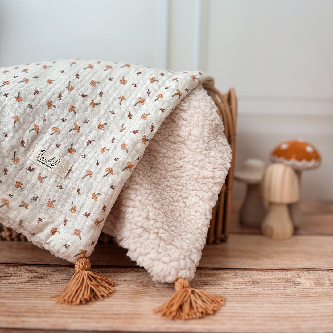 Découvrez la couverture pour bébé idéale pour apporter chaleur et douceur au quotidien de votre tout-petit ! Confectionnée à la main en France, cette couverture unique se compose d’une face au motif champignon enchanteur et d’une face en fausse fourrure sherpa beige extra douce, assurant confort et style. Pour un charme supplémentaire, quatre pompons couleur cassonade décorent élégamment chaque coin de la couverture. Et pour une touche encore plus personnelle, elle peut être brodée avec le prénom de votre b