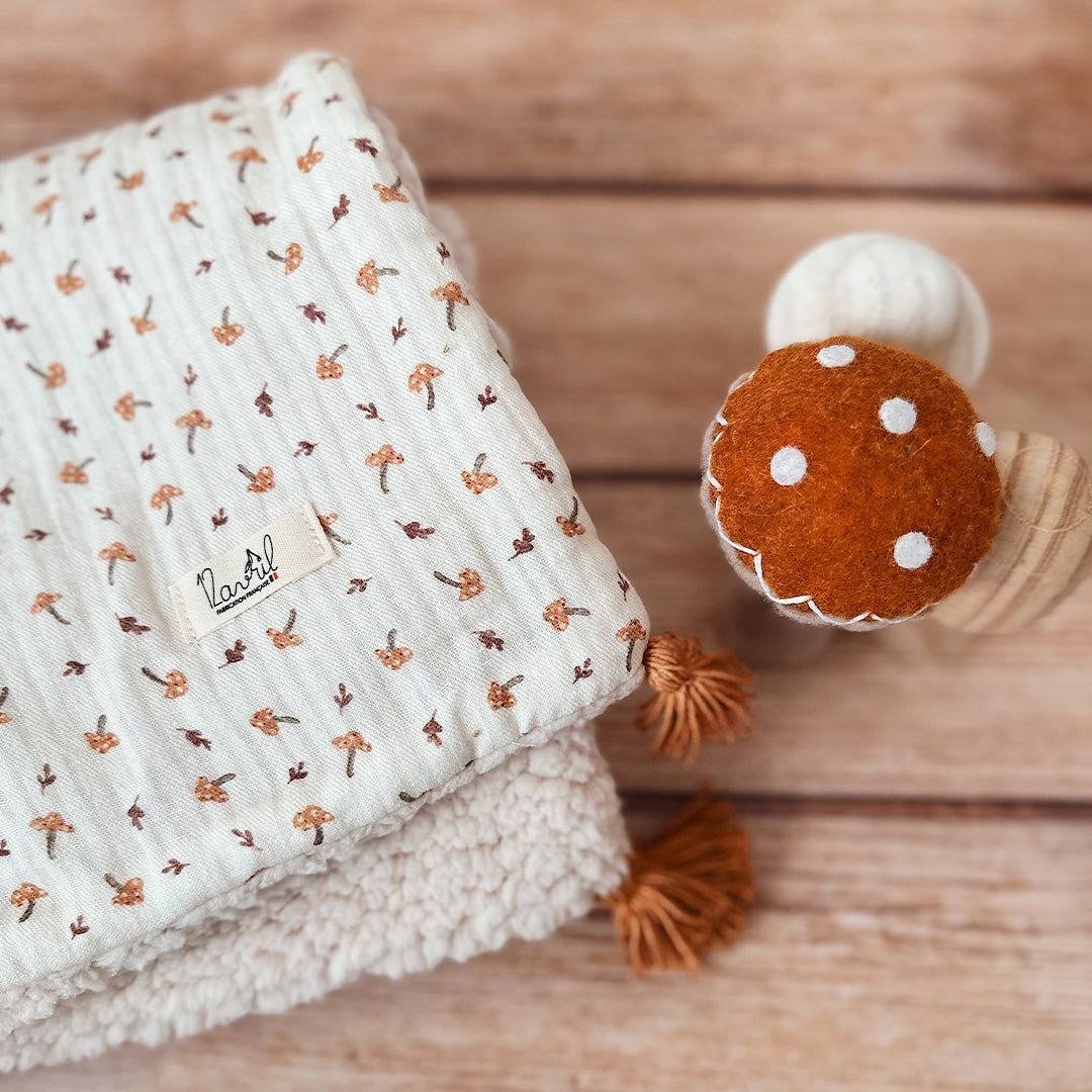 Découvrez la couverture pour bébé idéale pour apporter chaleur et douceur au quotidien de votre tout-petit ! Confectionnée à la main en France, cette couverture unique se compose d’une face au motif champignon enchanteur et d’une face en fausse fourrure sherpa beige extra douce, assurant confort et style. Pour un charme supplémentaire, quatre pompons couleur cassonade décorent élégamment chaque coin de la couverture. Et pour une touche encore plus personnelle, elle peut être brodée avec le prénom de votre b