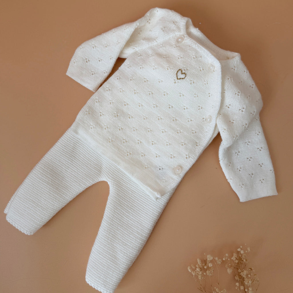 Offrez à votre bébé un premier vêtement doux, élégant et entièrement fabriqué en France avec cet adorable ensemble de naissance en maille blanche. Composé d'une brassière au col en dentelle de coton et d'un pantalon assorti, cet ensemble est tricoté avec soin dans une maille jersey, un point traditionnel qui lui confère un relief raffiné et un toucher délicatement texturé. La brassière croisée s'attache facilement grâce à de petits boutons  pour habiller bébé tout en douceur et sans effort. Le pantalon à ta