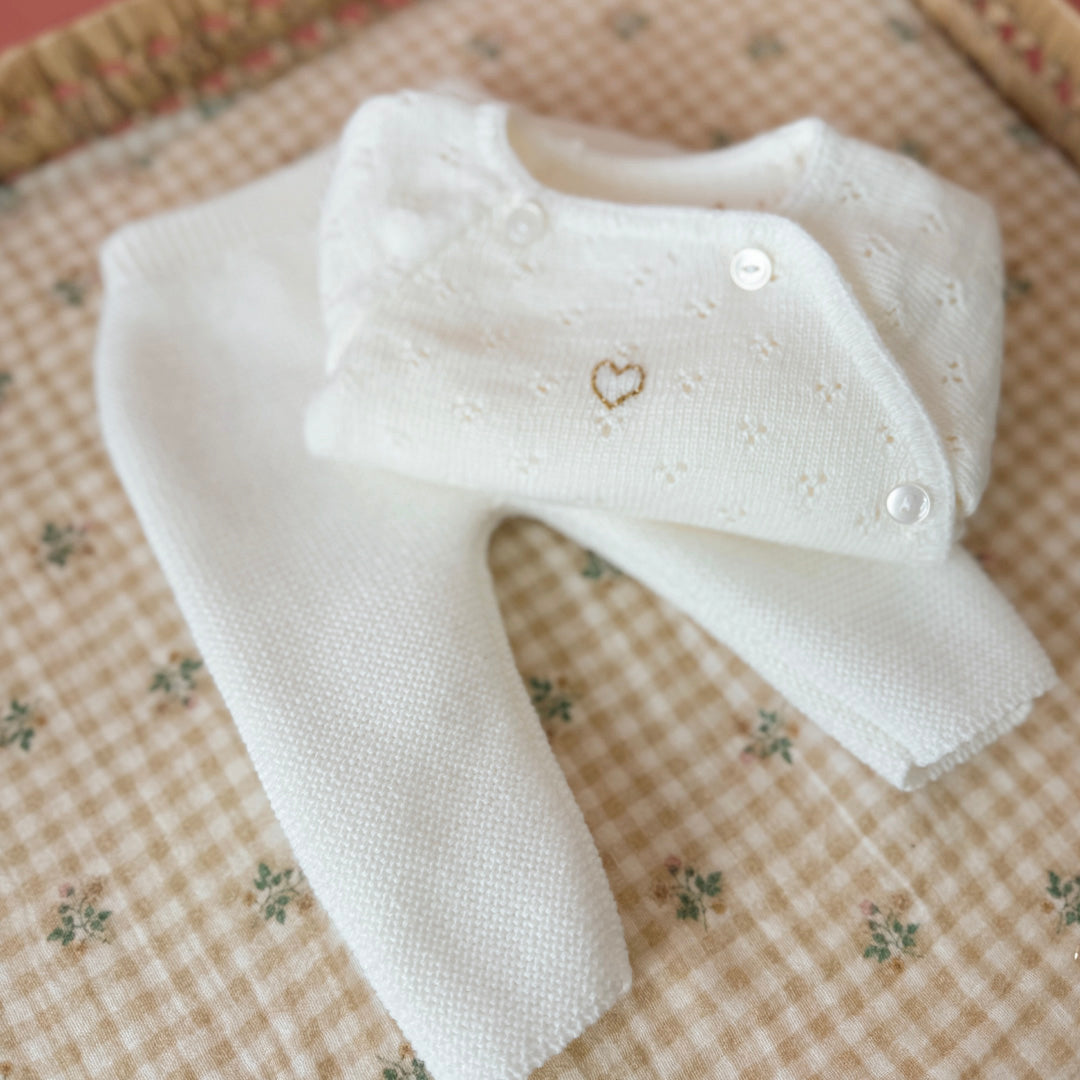 Offrez à votre bébé un premier vêtement doux, élégant et entièrement fabriqué en France avec cet adorable ensemble de naissance en maille blanche. Composé d'une brassière au col en dentelle de coton et d'un pantalon assorti, cet ensemble est tricoté avec soin dans une maille jersey, un point traditionnel qui lui confère un relief raffiné et un toucher délicatement texturé. La brassière croisée s'attache facilement grâce à de petits boutons  pour habiller bébé tout en douceur et sans effort. Le pantalon à ta
