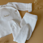 Offrez à votre bébé un premier vêtement doux, élégant et entièrement fabriqué en France avec cet adorable ensemble de naissance en maille blanche. Composé d'une brassière croisée et d'un pantalon assorti, cet ensemble est tricoté avec soin dans une maille jersey, un point traditionnel qui lui confère un relief raffiné et un toucher délicatement texturé. La brassière croisée s'attache facilement grâce à de petits boutons coloris bois, pour habiller bébé tout en douceur et sans effort. Le pantalon à taille él