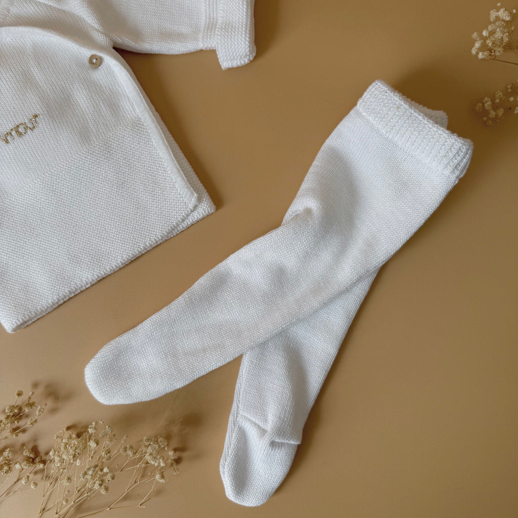 Offrez à votre bébé un premier vêtement doux, élégant et entièrement fabriqué en France avec cet adorable ensemble de naissance en maille blanche. Composé d'une brassière croisée et d'un pantalon assorti, cet ensemble est tricoté avec soin dans une maille jersey, un point traditionnel qui lui confère un relief raffiné et un toucher délicatement texturé. La brassière croisée s'attache facilement grâce à de petits boutons coloris bois, pour habiller bébé tout en douceur et sans effort. Le pantalon à taille él