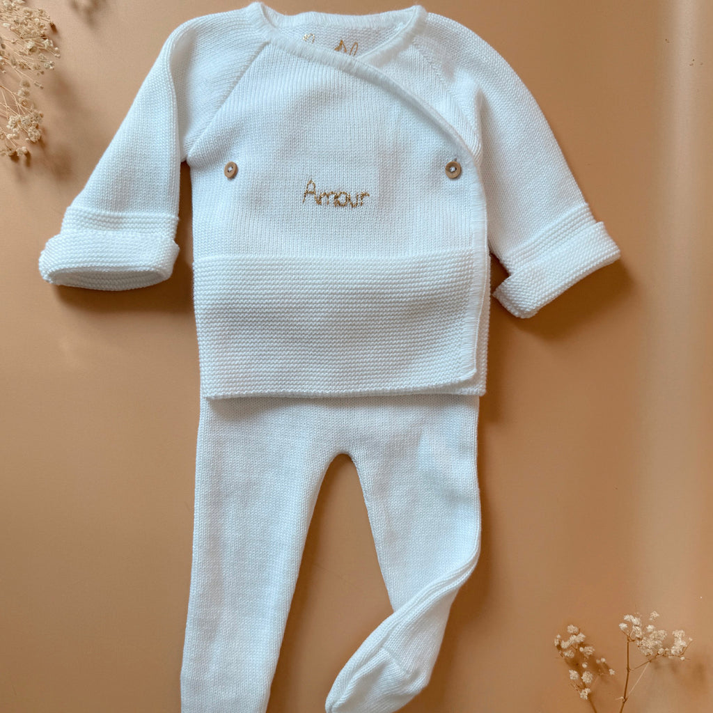 Offrez à votre bébé un premier vêtement doux, élégant et entièrement fabriqué en France avec cet adorable ensemble de naissance en maille blanche. Composé d'une brassière croisée et d'un pantalon assorti, cet ensemble est tricoté avec soin dans une maille jersey, un point traditionnel qui lui confère un relief raffiné et un toucher délicatement texturé. La brassière croisée s'attache facilement grâce à de petits boutons coloris bois, pour habiller bébé tout en douceur et sans effort. Le pantalon à taille él