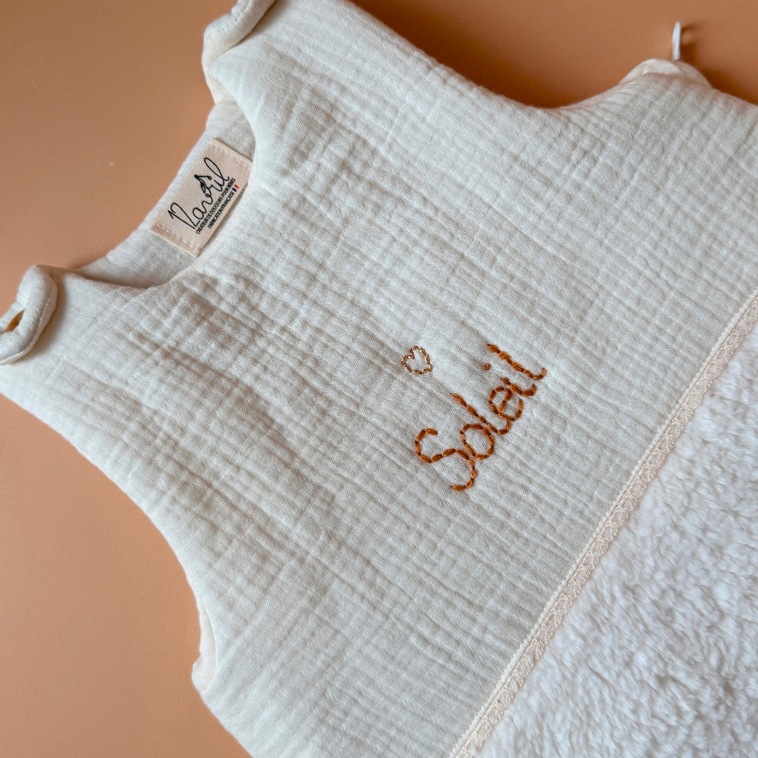 Cette gigoteuse pour bébé, confectionnée avec soin, est le choix idéal pour garder bébé au chaud durant l’hiver. Fabriquée en fausse fourrure type sherpa et en gaze de coton écrue, elle allie douceur et élégance. Son design raffiné, avec un empiècement en gaze de coton orné d'un discret galon en coton ajouré, en fait un accessoire de choix pour un sommeil cosy et stylé. Personnalisez la gigoteuse de bébé Vous avez la possibilité de personnaliser la gigoteuse avec le prénom ou le surnom de bébé, brodé à la m