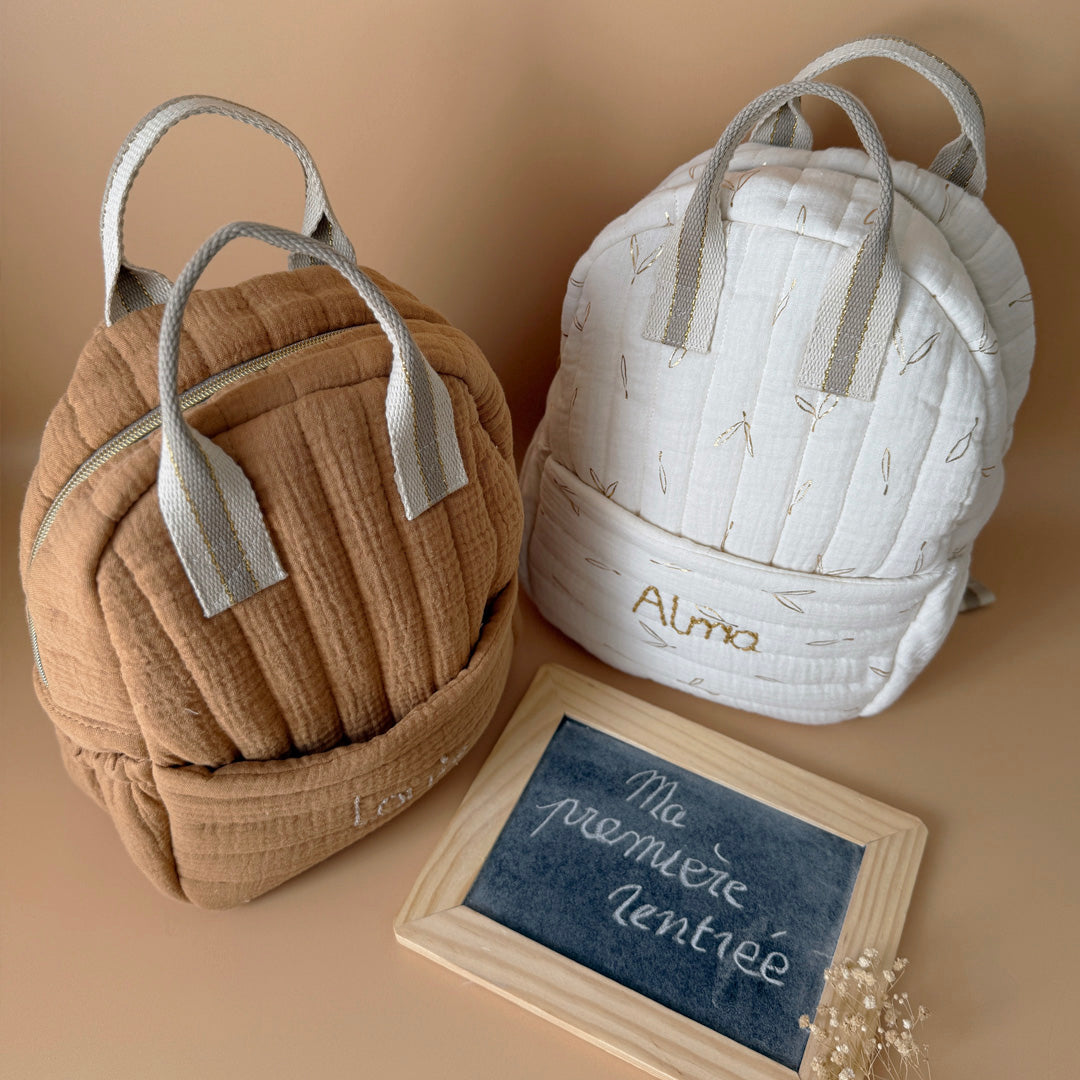 Découvrez notre sac à dos maternelle personnalisé, confectionné avec soin en France pour accompagner les tout-petits dans leurs premières aventures à l’école. Ce modèle est réalisé en gaze de coton matelassé, douce et respirante, dans un joli coloris biscuit chaleureux et intemporel. Ce sac se distingue par sa personnalisation brodée à la main : le prénom de l’enfant est délicatement inscrit sur la poche avant avec un fil doré clair ou argenté, pour un rendu raffiné et unique. Une attention particulière qui