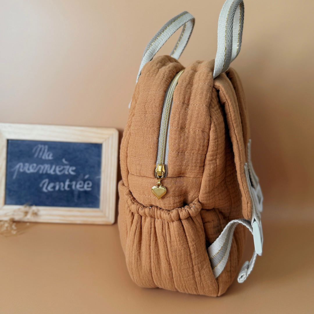 Découvrez notre sac à dos maternelle personnalisé, confectionné avec soin en France pour accompagner les tout-petits dans leurs premières aventures à l’école. Ce modèle est réalisé en gaze de coton matelassé, douce et respirante, dans un joli coloris biscuit chaleureux et intemporel. Ce sac se distingue par sa personnalisation brodée à la main : le prénom de l’enfant est délicatement inscrit sur la poche avant avec un fil doré clair ou argenté, pour un rendu raffiné et unique. Une attention particulière qui