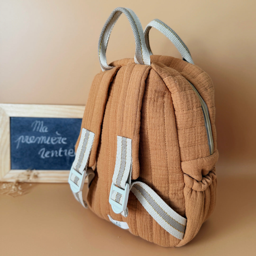 Découvrez notre sac à dos maternelle personnalisé, confectionné avec soin en France pour accompagner les tout-petits dans leurs premières aventures à l’école. Ce modèle est réalisé en gaze de coton matelassé, douce et respirante, dans un joli coloris biscuit chaleureux et intemporel. Ce sac se distingue par sa personnalisation brodée à la main : le prénom de l’enfant est délicatement inscrit sur la poche avant avec un fil doré clair ou argenté, pour un rendu raffiné et unique. Une attention particulière qui