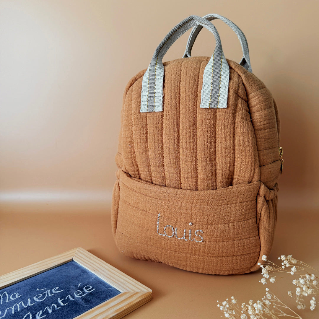 Découvrez notre sac à dos maternelle personnalisé, confectionné avec soin en France pour accompagner les tout-petits dans leurs premières aventures à l’école. Ce modèle est réalisé en gaze de coton matelassé, douce et respirante, dans un joli coloris biscuit chaleureux et intemporel. Ce sac se distingue par sa personnalisation brodée à la main : le prénom de l’enfant est délicatement inscrit sur la poche avant avec un fil doré clair ou argenté, pour un rendu raffiné et unique. Une attention particulière qui