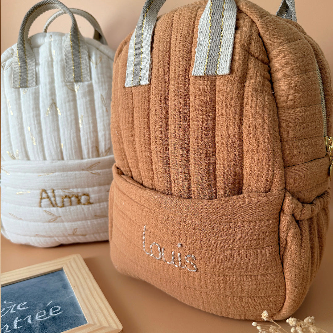 Découvrez notre sac à dos maternelle personnalisé, confectionné avec soin en France pour accompagner les tout-petits dans leurs premières aventures à l’école. Ce modèle est réalisé en gaze de coton matelassé, douce et respirante, dans un joli coloris biscuit chaleureux et intemporel. Ce sac se distingue par sa personnalisation brodée à la main : le prénom de l’enfant est délicatement inscrit sur la poche avant avec un fil doré clair ou argenté, pour un rendu raffiné et unique. Une attention particulière qui