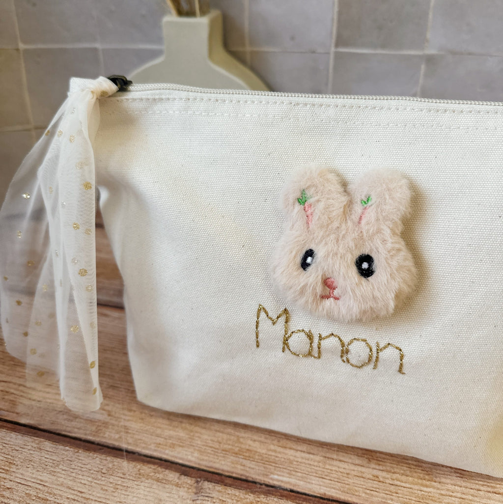 Cette jolie trousse de toilette deviendra vite l' indispensable de bébé à glisser partout ! Elle est réalisée en 100% toile de coton biologique. Une jolie petite tête de lapin en fourrure est délicatement cousue , puis nous brodons le prénom que vous désirez avec un fil de lurex doré. Le tout est contrecollé à l'intérieur pour un maximum de solidité. Son ouverture zippée est agrémentée par un lien en tulle à pois doré, ce qui facilitera l'ouverture.