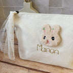 Cette jolie trousse de toilette deviendra vite l' indispensable de bébé à glisser partout ! Elle est réalisée en 100% toile de coton biologique. Une jolie petite tête de lapin en fourrure est délicatement cousue , puis nous brodons le prénom que vous désirez avec un fil de lurex doré. Le tout est contrecollé à l'intérieur pour un maximum de solidité. Son ouverture zippée est agrémentée par un lien en tulle à pois doré, ce qui facilitera l'ouverture.