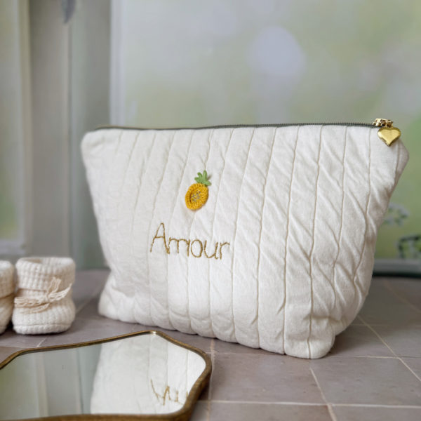 Apportez une touche de tendresse à vos accessoires avec cette trousse de toilette matelassée en jersey de coton, entièrement confectionnée en France. Pensée pour les mamans, les enfants ou tout simplement pour se faire plaisir, cette trousse incarne à la fois le charme artisanal et la fonctionnalité. Son tissu matelassé lui confère une allure douce et moelleuse, parfaite pour y glisser les essentiels de toilette ou les petits trésors du quotidien. Ce qui la rend unique ? Son joli petit ananas jaune mordoré cousu sur le devant, associée à une broderie  personnalisée au prénom de votre choix. Chaque trousse est brodée à la main avec soin, rendant chaque pièce unique et affectueuse. Idéale comme cadeau de naissance, de babyshower, ou pour toute occasion où l’on veut faire plaisir avec du fait-main de qualité. Fièrement fabriquée en France, cette trousse de toilette est le fruit d'un savoir-faire artisanal et d'une attention particulière aux détails. Zoom sur le petit curseur de la fermeture à glissière !