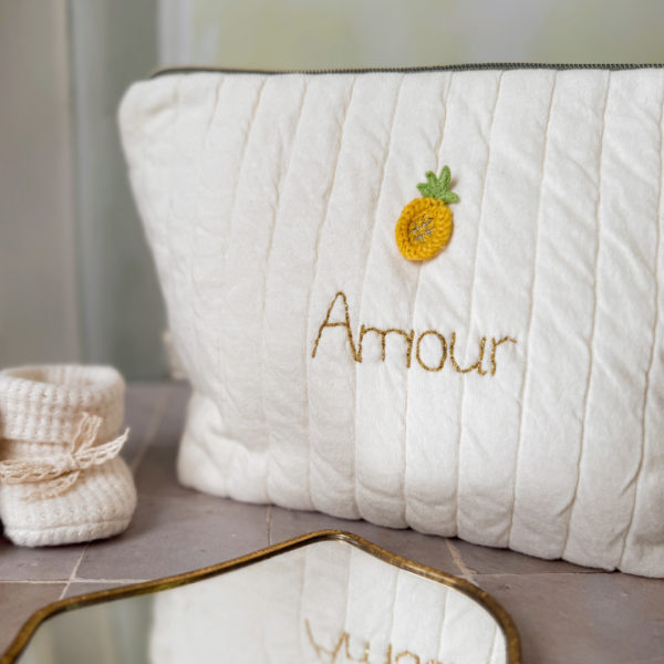 Apportez une touche de tendresse à vos accessoires avec cette trousse de toilette matelassée en jersey de coton, entièrement confectionnée en France. Pensée pour les mamans, les enfants ou tout simplement pour se faire plaisir, cette trousse incarne à la fois le charme artisanal et la fonctionnalité. Son tissu matelassé lui confère une allure douce et moelleuse, parfaite pour y glisser les essentiels de toilette ou les petits trésors du quotidien. Ce qui la rend unique ? Son joli petit ananas jaune mordoré cousu sur le devant, associée à une broderie  personnalisée au prénom de votre choix. Chaque trousse est brodée à la main avec soin, rendant chaque pièce unique et affectueuse. Idéale comme cadeau de naissance, de babyshower, ou pour toute occasion où l’on veut faire plaisir avec du fait-main de qualité. Fièrement fabriquée en France, cette trousse de toilette est le fruit d'un savoir-faire artisanal et d'une attention particulière aux détails. Zoom sur le petit curseur de la fermeture à glissière !