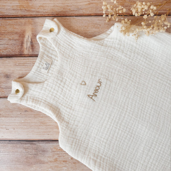 Gigoteuse d'Été en Triple Gaze de Coton Bio – Personnalisable Offrez à votre bébé un confort inégalé pendant les nuits d'été avec cette magnifique gigoteuse confectionnée dans une triple gaze de coton biologique certifiée Oeko-Tex, une matière d'exception bien plus douce et respirante que la traditionnelle double gaze. Sa texture gaufrée naturelle apporte douceur, légèreté et élégance. Disponible en coloris écru, cette gigoteuse est personnalisable au prénom de votre enfant, délicatement brodé sous un petit cœur doré au centre, ajoutant une touche poétique et unique. Conçue pour faciliter le quotidien des parents, elle s'ouvre aisément grâce à deux pressions dorées aux épaules et une grande fermeture à glissière latérale, permettant un habillage simple et rapide, même lorsque bébé dort. Un véritable cocon de tendresse, pensé pour le bien-être des tout-petits et la tranquillité des grands.