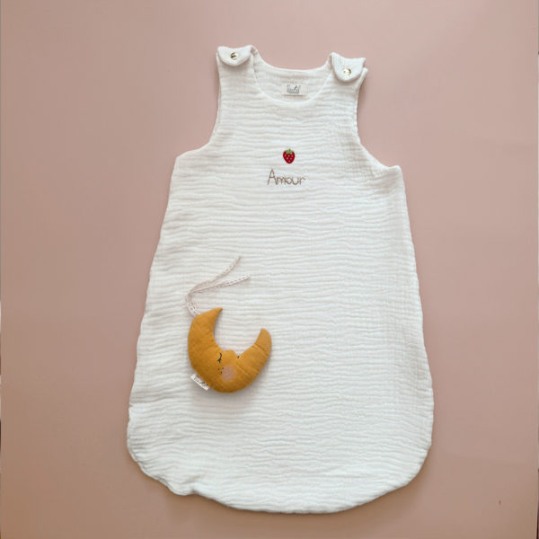 ffrez à votre bébé un confort inégalé pendant les nuits d'été avec cette magnifique gigoteuse confectionnée dans une triple gaze de coton biologique certifiée Oeko-Tex, une matière d'exception bien plus douce et respirante que la traditionnelle double gaze. Sa texture gaufrée naturelle apporte douceur, légèreté et élégance. Disponible en coloris écru, cette gigoteuse est personnalisable au prénom de votre enfant, délicatement brodé sous une petite fraise appliquée au centre, ajoutant une touche fruitée et unique. Conçue pour faciliter le quotidien des parents, elle s'ouvre aisément grâce à deux pressions dorées aux épaules et une grande fermeture à glissière latérale, permettant un habillage simple et rapide, même lorsque bébé dort. Un véritable cocon de tendresse, pensé pour le bien-être des tout-petits et la tranquillité des grands. Le petit détail qui fait toute la différence : la gigoteuse est personnalisable avec le prénom de votre bébé, brodé à la main dans un joli fil doré, pour un rendu raffiné et unique. Juste au-dessus, une petite fraise appliquée et brodée apporte une touche estivale pleine de fraîcheur et de poésie. Facile à enfiler, elle s’ouvre par deux pressions dorées aux épaules et une grande fermeture zippée sur le côté, pour un habillage rapide, même pendant le sommeil.