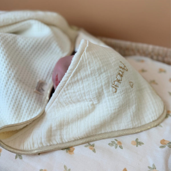 Offrez à votre nouveau-né une sortie de bain tout en douceur avec notre cape de bain de naissance en nid d’éponge, confectionnée en coton 100% naturel. Cette cape innovante associe deux textures complémentaires : L’éponge ultra absorbante, idéale pour sécher bébé rapidement, Et le nid d’abeille moelleux, parfait pour envelopper délicatement sa peau fragile. Sa forme carrée de 70 x 70 cm est spécialement pensée pour les tout-petits. Elle épouse parfaitement la taille de bébé pendant ses premiers mois, sans excès de tissu, et permet une manipulation facile après le bain. La capuche en gaze de coton, douce et respirante, protège sa petite tête. En option, vous pouvez la faire broder au prénom de votre bébé, pour un souvenir unique et plein d’émotion. Une broderie délicate accompagnée d’un petit cœur doré ajoute une touche de tendresse et d’élégance à l’ensemble. Chaque cape est gansée d’un biais beige doré, pour une finition soignée et raffinée qui en fait un véritable objet de naissance haut de gamme.