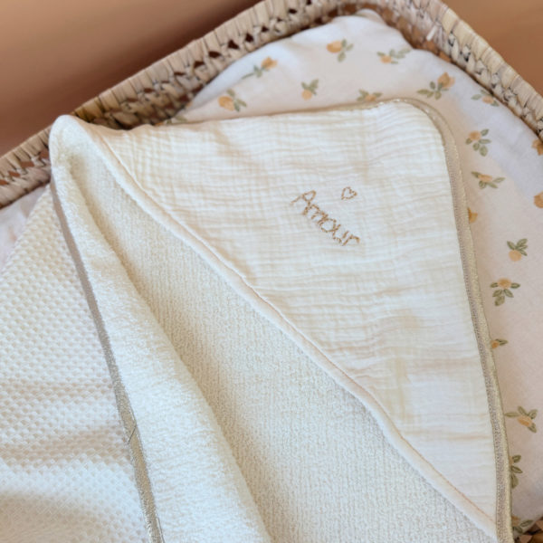 Offrez à votre nouveau-né une sortie de bain tout en douceur avec notre cape de bain de naissance en nid d’éponge, confectionnée en coton 100% naturel. Cette cape innovante associe deux textures complémentaires : L’éponge ultra absorbante, idéale pour sécher bébé rapidement, Et le nid d’abeille moelleux, parfait pour envelopper délicatement sa peau fragile. Sa forme carrée de 70 x 70 cm est spécialement pensée pour les tout-petits. Elle épouse parfaitement la taille de bébé pendant ses premiers mois, sans excès de tissu, et permet une manipulation facile après le bain. La capuche en gaze de coton, douce et respirante, protège sa petite tête. En option, vous pouvez la faire broder au prénom de votre bébé, pour un souvenir unique et plein d’émotion. Une broderie délicate accompagnée d’un petit cœur doré ajoute une touche de tendresse et d’élégance à l’ensemble. Chaque cape est gansée d’un biais beige doré, pour une finition soignée et raffinée qui en fait un véritable objet de naissance haut de gamme.