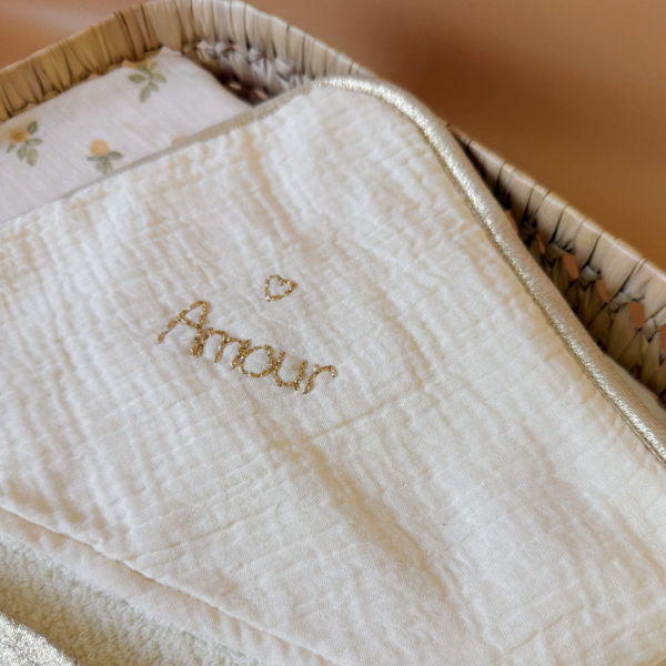 Offrez à votre nouveau-né une sortie de bain tout en douceur avec notre cape de bain de naissance en nid d’éponge, confectionnée en coton 100% naturel. Cette cape innovante associe deux textures complémentaires : L’éponge ultra absorbante, idéale pour sécher bébé rapidement, Et le nid d’abeille moelleux, parfait pour envelopper délicatement sa peau fragile. Sa forme carrée de 70 x 70 cm est spécialement pensée pour les tout-petits. Elle épouse parfaitement la taille de bébé pendant ses premiers mois, sans excès de tissu, et permet une manipulation facile après le bain. La capuche en gaze de coton, douce et respirante, protège sa petite tête. En option, vous pouvez la faire broder au prénom de votre bébé, pour un souvenir unique et plein d’émotion. Une broderie délicate accompagnée d’un petit cœur doré ajoute une touche de tendresse et d’élégance à l’ensemble. Chaque cape est gansée d’un biais beige doré, pour une finition soignée et raffinée qui en fait un véritable objet de naissance haut de gamme.
