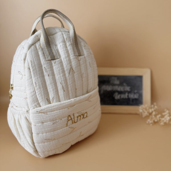 Voici notre tout nouveau sac à dos maternelle, pensé et confectionné en France pour accompagner vos tout-petits dans leurs premières journées d'école. Ce modèle délicat est réalisé en gaze de coton matelassée écrue, ornée de petites feuilles dorées qui lui donnent une allure douce et poétique. La grande particularité de ce sac ? Il est personnalisé avec le prénom de votre enfant, brodé à la main en fil doré sur la poche avant. Un détail unique, raffiné, et tellement précieux pour marquer les affaires de votre petit. L’intérieur du sac est entièrement doublé avec un tissu imperméable, parfait pour protéger son contenu contre les petits accidents ou les surprises météo. Son ouverture zippée large a été pensée pour que les enfants puissent l’utiliser en toute autonomie. Il comporte trois poches intelligentes, dont deux latérales élastiquées idéales pour glisser une gourde, un doudou ou les trésors ramassés à la récréation. Côté confort, rien n’a été laissé au hasard : bretelles matelassées réglables et ajustables pour un bon maintien sans tirer sur les épaules, et deux anses solides pour une prise en main facile par les petits ou les adultes. C'est plus qu’un sac à dos : c’est un vrai compagnon de rentrée, beau, pratique, doux pour le dos, et surtout unique grâce à sa personnalisation.
