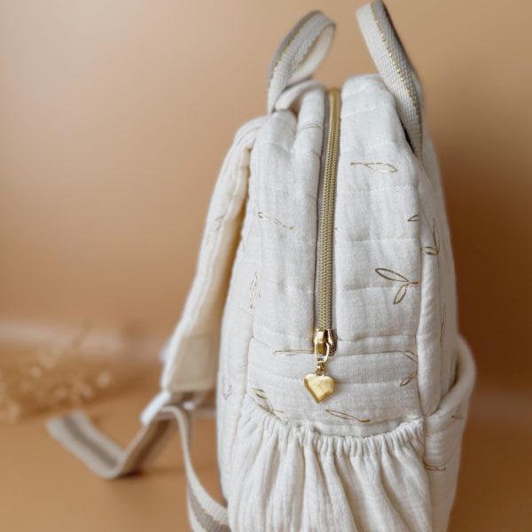 Voici notre tout nouveau sac à dos maternelle, pensé et confectionné en France pour accompagner vos tout-petits dans leurs premières journées d'école. Ce modèle délicat est réalisé en gaze de coton matelassée écrue, ornée de petites feuilles dorées qui lui donnent une allure douce et poétique. La grande particularité de ce sac ? Il est personnalisé avec le prénom de votre enfant, brodé à la main en fil doré sur la poche avant. Un détail unique, raffiné, et tellement précieux pour marquer les affaires de votre petit. L’intérieur du sac est entièrement doublé avec un tissu imperméable, parfait pour protéger son contenu contre les petits accidents ou les surprises météo. Son ouverture zippée large a été pensée pour que les enfants puissent l’utiliser en toute autonomie. Il comporte trois poches intelligentes, dont deux latérales élastiquées idéales pour glisser une gourde, un doudou ou les trésors ramassés à la récréation. Côté confort, rien n’a été laissé au hasard : bretelles matelassées réglables et ajustables pour un bon maintien sans tirer sur les épaules, et deux anses solides pour une prise en main facile par les petits ou les adultes. C'est plus qu’un sac à dos : c’est un vrai compagnon de rentrée, beau, pratique, doux pour le dos, et surtout unique grâce à sa personnalisation.