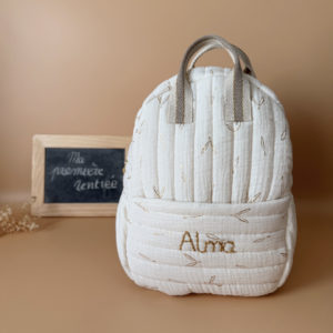 Voici notre tout nouveau sac à dos maternelle, pensé et confectionné en France pour accompagner vos tout-petits dans leurs premières journées d'école. Ce modèle délicat est réalisé en gaze de coton matelassée écrue, ornée de petites feuilles dorées qui lui donnent une allure douce et poétique. La grande particularité de ce sac ? Il est personnalisé avec le prénom de votre enfant, brodé à la main en fil doré sur la poche avant. Un détail unique, raffiné, et tellement précieux pour marquer les affaires de votre petit. L’intérieur du sac est entièrement doublé avec un tissu imperméable, parfait pour protéger son contenu contre les petits accidents ou les surprises météo. Son ouverture zippée large a été pensée pour que les enfants puissent l’utiliser en toute autonomie. Il comporte trois poches intelligentes, dont deux latérales élastiquées idéales pour glisser une gourde, un doudou ou les trésors ramassés à la récréation. Côté confort, rien n’a été laissé au hasard : bretelles matelassées réglables et ajustables pour un bon maintien sans tirer sur les épaules, et deux anses solides pour une prise en main facile par les petits ou les adultes. C'est plus qu’un sac à dos : c’est un vrai compagnon de rentrée, beau, pratique, doux pour le dos, et surtout unique grâce à sa personnalisation.