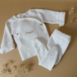 Offrez à votre bébé un premier vêtement doux, élégant et entièrement fabriqué en France avec cet adorable ensemble de naissance en maille blanche. Composé d'une brassière croisée et d'un pantalon assorti, cet ensemble est tricoté avec soin dans une maille jersey, un point traditionnel qui lui confère un relief raffiné et un toucher délicatement texturé. La brassière croisée s'attache facilement grâce à de petits boutons coloris bois, pour habiller bébé tout en douceur et sans effort. Le pantalon à taille élastiquée et à pieds  assure un confort optimal et un bon maintien, sans comprimer le ventre fragile des nouveau-nés. Ce modèle est intégralement tricoté en France, en collaboration avec un atelier partenaire reconnu pour son savoir-faire en maille fine pour bébé. Une attention toute particulière est portée à la qualité des finitions, pour un rendu chic, intemporel et parfaitement adapté à la peau des tout-petits. Et pour rendre ce cadeau de naissance encore plus unique, vous pouvez choisir de personnaliser la brassière avec une broderie faite main, directement au centre du vêtement. Que ce soit le prénom du bébé ou un petit mot doux, chaque pièce devient ainsi un souvenir précieux, plein d’émotion.
