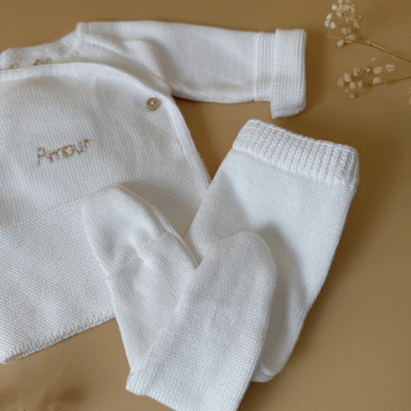 Offrez à votre bébé un premier vêtement doux, élégant et entièrement fabriqué en France avec cet adorable ensemble de naissance en maille blanche. Composé d'une brassière croisée et d'un pantalon assorti, cet ensemble est tricoté avec soin dans une maille jersey, un point traditionnel qui lui confère un relief raffiné et un toucher délicatement texturé. La brassière croisée s'attache facilement grâce à de petits boutons coloris bois, pour habiller bébé tout en douceur et sans effort. Le pantalon à taille élastiquée et à pieds  assure un confort optimal et un bon maintien, sans comprimer le ventre fragile des nouveau-nés. Ce modèle est intégralement tricoté en France, en collaboration avec un atelier partenaire reconnu pour son savoir-faire en maille fine pour bébé. Une attention toute particulière est portée à la qualité des finitions, pour un rendu chic, intemporel et parfaitement adapté à la peau des tout-petits. Et pour rendre ce cadeau de naissance encore plus unique, vous pouvez choisir de personnaliser la brassière avec une broderie faite main, directement au centre du vêtement. Que ce soit le prénom du bébé ou un petit mot doux, chaque pièce devient ainsi un souvenir précieux, plein d’émotion.