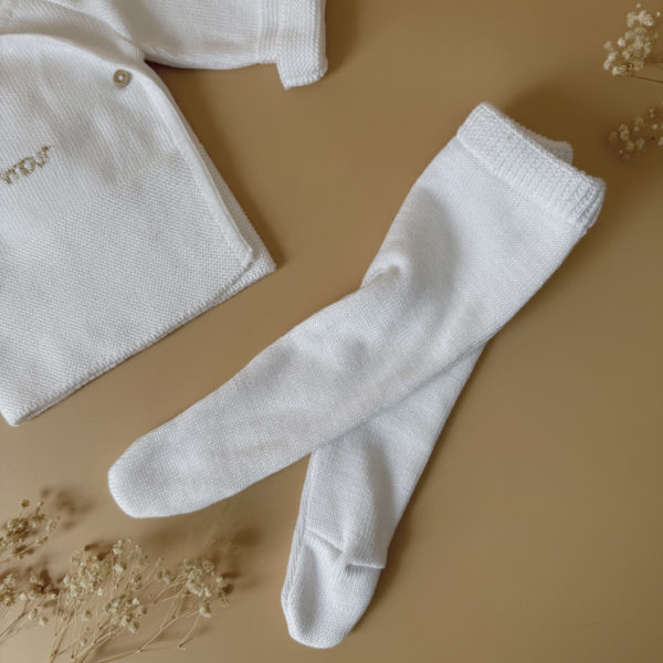 Offrez à votre bébé un premier vêtement doux, élégant et entièrement fabriqué en France avec cet adorable ensemble de naissance en maille blanche. Composé d'une brassière croisée et d'un pantalon assorti, cet ensemble est tricoté avec soin dans une maille jersey, un point traditionnel qui lui confère un relief raffiné et un toucher délicatement texturé. La brassière croisée s'attache facilement grâce à de petits boutons coloris bois, pour habiller bébé tout en douceur et sans effort. Le pantalon à taille élastiquée et à pieds  assure un confort optimal et un bon maintien, sans comprimer le ventre fragile des nouveau-nés. Ce modèle est intégralement tricoté en France, en collaboration avec un atelier partenaire reconnu pour son savoir-faire en maille fine pour bébé. Une attention toute particulière est portée à la qualité des finitions, pour un rendu chic, intemporel et parfaitement adapté à la peau des tout-petits. Et pour rendre ce cadeau de naissance encore plus unique, vous pouvez choisir de personnaliser la brassière avec une broderie faite main, directement au centre du vêtement. Que ce soit le prénom du bébé ou un petit mot doux, chaque pièce devient ainsi un souvenir précieux, plein d’émotion.