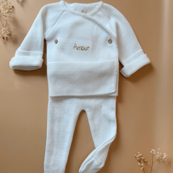 Offrez à votre bébé un premier vêtement doux, élégant et entièrement fabriqué en France avec cet adorable ensemble de naissance en maille blanche. Composé d'une brassière croisée et d'un pantalon assorti, cet ensemble est tricoté avec soin dans une maille jersey, un point traditionnel qui lui confère un relief raffiné et un toucher délicatement texturé. La brassière croisée s'attache facilement grâce à de petits boutons coloris bois, pour habiller bébé tout en douceur et sans effort. Le pantalon à taille élastiquée et à pieds  assure un confort optimal et un bon maintien, sans comprimer le ventre fragile des nouveau-nés. Ce modèle est intégralement tricoté en France, en collaboration avec un atelier partenaire reconnu pour son savoir-faire en maille fine pour bébé. Une attention toute particulière est portée à la qualité des finitions, pour un rendu chic, intemporel et parfaitement adapté à la peau des tout-petits. Et pour rendre ce cadeau de naissance encore plus unique, vous pouvez choisir de personnaliser la brassière avec une broderie faite main, directement au centre du vêtement. Que ce soit le prénom du bébé ou un petit mot doux, chaque pièce devient ainsi un souvenir précieux, plein d’émotion.