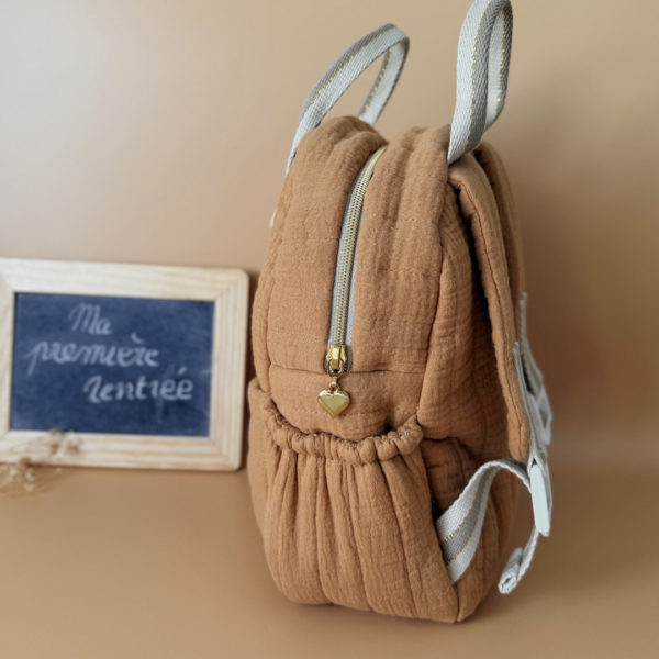 Découvrez notre sac à dos maternelle personnalisé, confectionné avec soin en France pour accompagner les tout-petits dans leurs premières aventures à l’école. Ce modèle est réalisé en gaze de coton matelassé, douce et respirante, dans un joli coloris biscuit chaleureux et intemporel. Ce sac se distingue par sa personnalisation brodée à la main : le prénom de l’enfant est délicatement inscrit sur la poche avant avec un fil doré clair ou argenté, pour un rendu raffiné et unique. Une attention particulière qui en fait un objet à la fois pratique et précieux. L’intérieur est entièrement doublé d’un tissu imperméable, afin de protéger le contenu des petits accidents du quotidien (gourdes mal fermées, goûter renversé, etc.). L’ouverture zippée est facile à manipuler par les petites mains, encourageant l’autonomie dès la petite section. Le sac est pensé pour être fonctionnel et ergonomique : il dispose de trois poches, dont deux latérales élastiquées idéales pour ranger une gourde, un doudou ou des petits jouets. Les bretelles matelassées et réglables assurent un confort optimal pour les épaules et le dos de votre enfant. Deux anses solides en sangle permettent de le porter facilement à la main. Ce sac à dos allie esthétique, confort et praticité, pour une rentrée maternelle sereine, tout en douceur.