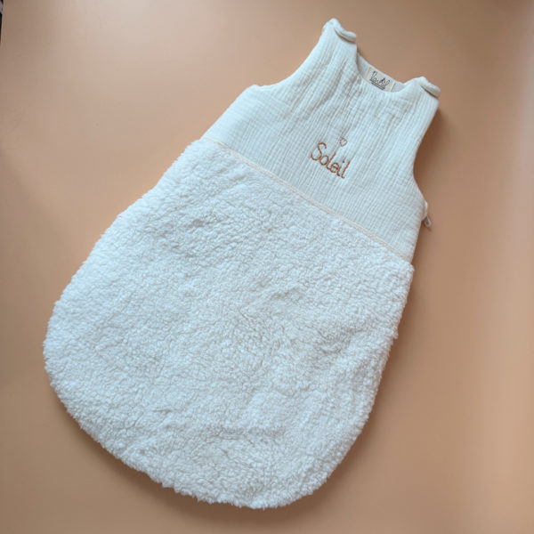 Cette gigoteuse pour bébé, confectionnée avec soin, est le choix idéal pour garder bébé au chaud durant l’hiver. Fabriquée en fausse fourrure type sherpa et en gaze de coton écrue, elle allie douceur et élégance. Son design raffiné, avec un empiècement en gaze de coton orné d'un discret galon en coton ajouré, en fait un accessoire de choix pour un sommeil cosy et stylé. Personnalisez la gigoteuse de bébé Vous avez la possibilité de personnaliser la gigoteuse avec le prénom ou le surnom de bébé, brodé à la main de la couleur de votre choix. La broderie est effectuée sur le milieu devant de la gigoteuse. Cette gigoteuse est parfaite pour : Garantir un sommeil chaud et confortable lors des nuits fraîches de l'hiver. Apporter une touche de raffinement à la chambre de bébé grâce à son design élégant. Offrir un cadeau de naissance pratique et raffiné. Avantages : Équivalent TOG 3 pour des chambres entre 18°C et 22°C Chaleur et confort : La fausse fourrure sherpa combinée à la doublure ouatinée apporte un confort thermique maximal. Élégance intemporelle : La broderie anglaise et l’empiècement en gaze de coton ajoutent un détail chic à cette gigoteuse. Pratique : Facile à enfiler et sécurisée, elle garantit un sommeil paisible sans risque de déperdition de chaleur.
