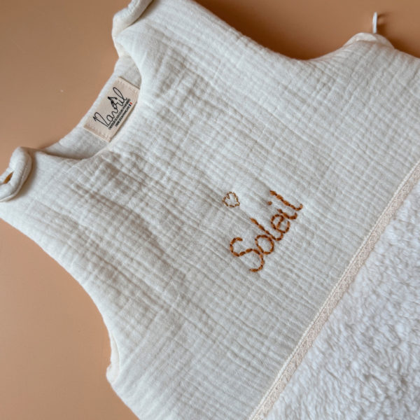 Cette gigoteuse pour bébé, confectionnée avec soin, est le choix idéal pour garder bébé au chaud durant l’hiver. Fabriquée en fausse fourrure type sherpa et en gaze de coton écrue, elle allie douceur et élégance. Son design raffiné, avec un empiècement en gaze de coton orné d'un discret galon en coton ajouré, en fait un accessoire de choix pour un sommeil cosy et stylé. Personnalisez la gigoteuse de bébé Vous avez la possibilité de personnaliser la gigoteuse avec le prénom ou le surnom de bébé, brodé à la main de la couleur de votre choix. La broderie est effectuée sur le milieu devant de la gigoteuse. Cette gigoteuse est parfaite pour : Garantir un sommeil chaud et confortable lors des nuits fraîches de l'hiver. Apporter une touche de raffinement à la chambre de bébé grâce à son design élégant. Offrir un cadeau de naissance pratique et raffiné. Avantages : Équivalent TOG 3 pour des chambres entre 18°C et 22°C Chaleur et confort : La fausse fourrure sherpa combinée à la doublure ouatinée apporte un confort thermique maximal. Élégance intemporelle : La broderie anglaise et l’empiècement en gaze de coton ajoutent un détail chic à cette gigoteuse. Pratique : Facile à enfiler et sécurisée, elle garantit un sommeil paisible sans risque de déperdition de chaleur.