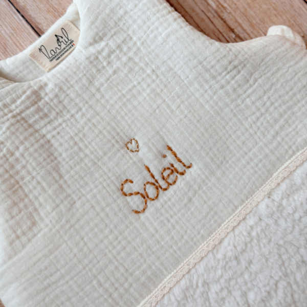 Cette gigoteuse pour bébé, confectionnée avec soin, est le choix idéal pour garder bébé au chaud durant l’hiver. Fabriquée en fausse fourrure type sherpa et en gaze de coton écrue, elle allie douceur et élégance. Son design raffiné, avec un empiècement en gaze de coton orné d'un discret galon en coton ajouré, en fait un accessoire de choix pour un sommeil cosy et stylé. Personnalisez la gigoteuse de bébé Vous avez la possibilité de personnaliser la gigoteuse avec le prénom ou le surnom de bébé, brodé à la main de la couleur de votre choix. La broderie est effectuée sur le milieu devant de la gigoteuse. Cette gigoteuse est parfaite pour : Garantir un sommeil chaud et confortable lors des nuits fraîches de l'hiver. Apporter une touche de raffinement à la chambre de bébé grâce à son design élégant. Offrir un cadeau de naissance pratique et raffiné. Avantages : Équivalent TOG 3 pour des chambres entre 18°C et 22°C Chaleur et confort : La fausse fourrure sherpa combinée à la doublure ouatinée apporte un confort thermique maximal. Élégance intemporelle : La broderie anglaise et l’empiècement en gaze de coton ajoutent un détail chic à cette gigoteuse. Pratique : Facile à enfiler et sécurisée, elle garantit un sommeil paisible sans risque de déperdition de chaleur.