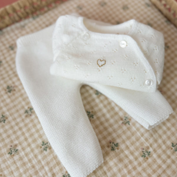 Offrez à votre bébé un premier vêtement doux, élégant et entièrement fabriqué en France avec cet adorable ensemble de naissance en maille blanche. Composé d'une brassière au col en dentelle de coton et d'un pantalon assorti, cet ensemble est tricoté avec soin dans une maille jersey, un point traditionnel qui lui confère un relief raffiné et un toucher délicatement texturé. La brassière croisée s'attache facilement grâce à de petits boutons  pour habiller bébé tout en douceur et sans effort. Le pantalon à taille élastiquée  assure un confort optimal et un bon maintien, sans comprimer le ventre fragile des nouveau-nés. Ce modèle est intégralement tricoté en France, en collaboration avec un atelier partenaire reconnu pour son savoir-faire en maille fine pour bébé. Une attention toute particulière est portée à la qualité des finitions, pour un rendu chic, intemporel et parfaitement adapté à la peau des tout-petits. Et pour rendre ce cadeau de naissance encore plus unique, vous pouvez choisir de personnaliser la brassière avec une broderie faite main, directement au centre du vêtement. Que ce soit le prénom du bébé ou un petit mot doux, chaque pièce devient ainsi un souvenir précieux, plein d’émotion.