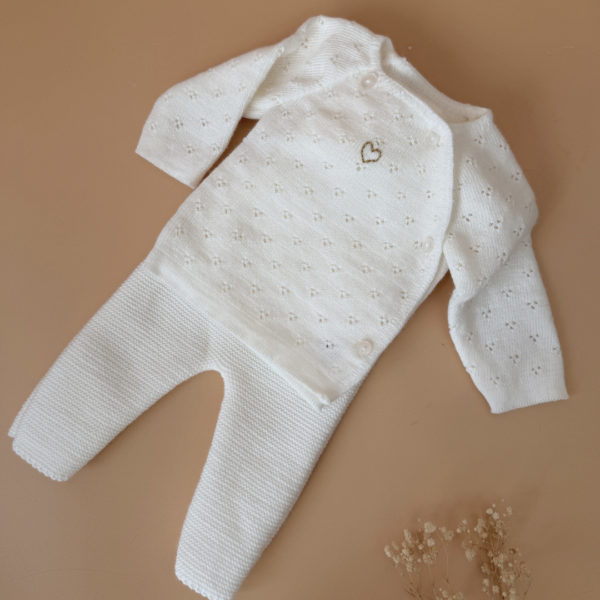 Offrez à votre bébé un premier vêtement doux, élégant et entièrement fabriqué en France avec cet adorable ensemble de naissance en maille blanche. Composé d'une brassière au col en dentelle de coton et d'un pantalon assorti, cet ensemble est tricoté avec soin dans une maille jersey, un point traditionnel qui lui confère un relief raffiné et un toucher délicatement texturé. La brassière croisée s'attache facilement grâce à de petits boutons  pour habiller bébé tout en douceur et sans effort. Le pantalon à taille élastiquée  assure un confort optimal et un bon maintien, sans comprimer le ventre fragile des nouveau-nés. Ce modèle est intégralement tricoté en France, en collaboration avec un atelier partenaire reconnu pour son savoir-faire en maille fine pour bébé. Une attention toute particulière est portée à la qualité des finitions, pour un rendu chic, intemporel et parfaitement adapté à la peau des tout-petits. Et pour rendre ce cadeau de naissance encore plus unique, vous pouvez choisir de personnaliser la brassière avec une broderie faite main, directement au centre du vêtement. Que ce soit le prénom du bébé ou un petit mot doux, chaque pièce devient ainsi un souvenir précieux, plein d’émotion.