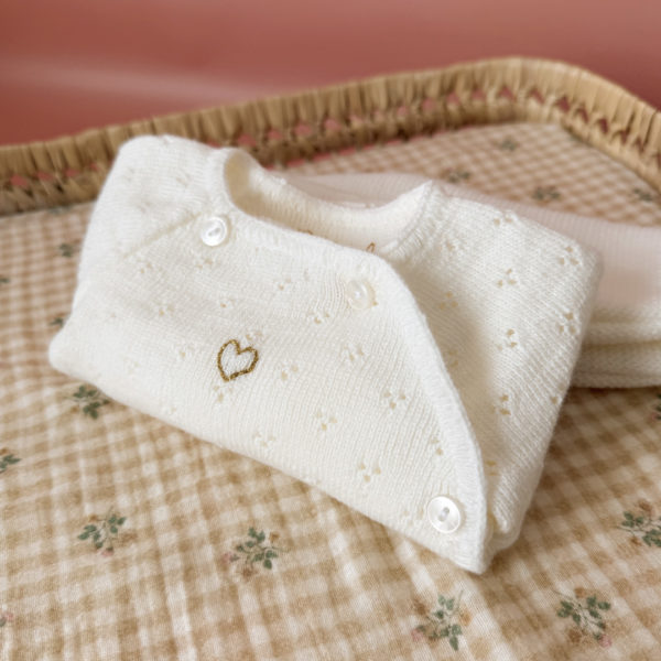 Offrez à votre bébé un premier vêtement doux, élégant et entièrement fabriqué en France avec cet adorable ensemble de naissance en maille blanche. Composé d'une brassière au col en dentelle de coton et d'un pantalon assorti, cet ensemble est tricoté avec soin dans une maille jersey, un point traditionnel qui lui confère un relief raffiné et un toucher délicatement texturé. La brassière croisée s'attache facilement grâce à de petits boutons  pour habiller bébé tout en douceur et sans effort. Le pantalon à taille élastiquée  assure un confort optimal et un bon maintien, sans comprimer le ventre fragile des nouveau-nés. Ce modèle est intégralement tricoté en France, en collaboration avec un atelier partenaire reconnu pour son savoir-faire en maille fine pour bébé. Une attention toute particulière est portée à la qualité des finitions, pour un rendu chic, intemporel et parfaitement adapté à la peau des tout-petits. Et pour rendre ce cadeau de naissance encore plus unique, vous pouvez choisir de personnaliser la brassière avec une broderie faite main, directement au centre du vêtement. Que ce soit le prénom du bébé ou un petit mot doux, chaque pièce devient ainsi un souvenir précieux, plein d’émotion.