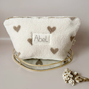 La trousse de toilette pour bébé est une pièce personnalisée et pratique, conçue avec soin pour répondre aux besoins des parents et de leur nouveau-né. Son grand format est très pratique.  La conception de la trousse permet aux produits de bébé de tenir debout à l'intérieur, ce qui facilite l'organisation et l'accès aux produits essentiels. Liniment, couches, lait de toilette, cotons, autant de produits pour un si petit bout de chou, que vous aurez besoin d'une grande trousse de toilette spacieuse. Fabriquée en fausse fourrure sherpa beige avec de jolis coeurs grège, cette trousse est à la fois douce et durable. L'intérieur de la trousse est doublé avec un tissu imperméable. Ls liquides renversés ne pénètrent pas à l'extérieur, protégeant ainsi les autres articles dans le sac à langer. Personnaliser votre trousse de toilette, au doux prénom de bébé ! La personnalisation est réalisée à la main sur le devant de la trousse de toilette avec un fil du coloris de votre choix.  C'est un plaston qui agrémente le devant de la trousse avec le prénom de bébé.  Fièrement fabriquée en France, cette trousse de toilette est le fruit d'un savoir-faire artisanal et d'une attention particulière aux détails. Zoom sur le petit curseur de la fermeture à glissière ! Découvrez notre gamme entière de produits autour du bain : de belles et grandes capes de bain XXL, langes et débarbouillettes. 