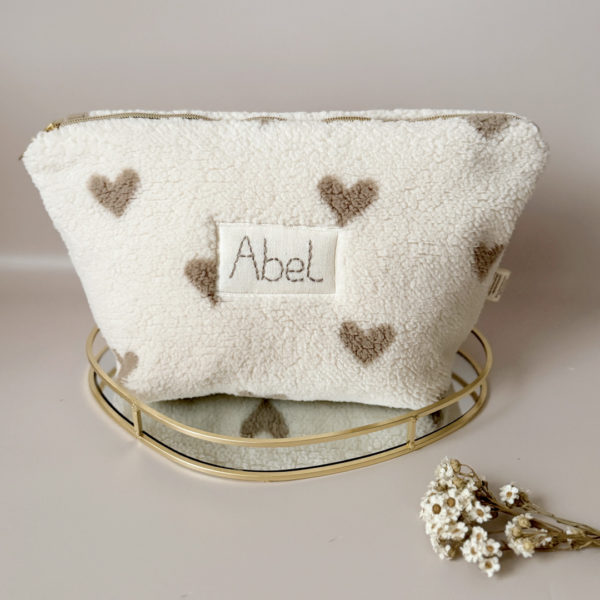 La trousse de toilette pour bébé est une pièce personnalisée et pratique, conçue avec soin pour répondre aux besoins des parents et de leur nouveau-né. Son grand format est très pratique.  La conception de la trousse permet aux produits de bébé de tenir debout à l'intérieur, ce qui facilite l'organisation et l'accès aux produits essentiels. Liniment, couches, lait de toilette, cotons, autant de produits pour un si petit bout de chou, que vous aurez besoin d'une grande trousse de toilette spacieuse. Fabriquée en fausse fourrure sherpa beige avec de jolis coeurs grège, cette trousse est à la fois douce et durable. L'intérieur de la trousse est doublé avec un tissu imperméable. Ls liquides renversés ne pénètrent pas à l'extérieur, protégeant ainsi les autres articles dans le sac à langer. Personnaliser votre trousse de toilette, au doux prénom de bébé ! La personnalisation est réalisée à la main sur le devant de la trousse de toilette avec un fil du coloris de votre choix.  C'est un plaston qui agrémente le devant de la trousse avec le prénom de bébé.  Fièrement fabriquée en France, cette trousse de toilette est le fruit d'un savoir-faire artisanal et d'une attention particulière aux détails. Zoom sur le petit curseur de la fermeture à glissière ! Découvrez notre gamme entière de produits autour du bain : de belles et grandes capes de bain XXL, langes et débarbouillettes. 