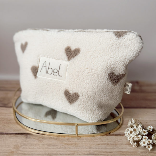 La trousse de toilette pour bébé est une pièce personnalisée et pratique, conçue avec soin pour répondre aux besoins des parents et de leur nouveau-né. Son grand format est très pratique.  La conception de la trousse permet aux produits de bébé de tenir debout à l'intérieur, ce qui facilite l'organisation et l'accès aux produits essentiels. Liniment, couches, lait de toilette, cotons, autant de produits pour un si petit bout de chou, que vous aurez besoin d'une grande trousse de toilette spacieuse. Fabriquée en fausse fourrure sherpa beige avec de jolis coeurs grège, cette trousse est à la fois douce et durable. L'intérieur de la trousse est doublé avec un tissu imperméable. Ls liquides renversés ne pénètrent pas à l'extérieur, protégeant ainsi les autres articles dans le sac à langer. Personnaliser votre trousse de toilette, au doux prénom de bébé ! La personnalisation est réalisée à la main sur le devant de la trousse de toilette avec un fil du coloris de votre choix.  C'est un plaston qui agrémente le devant de la trousse avec le prénom de bébé.  Fièrement fabriquée en France, cette trousse de toilette est le fruit d'un savoir-faire artisanal et d'une attention particulière aux détails. Zoom sur le petit curseur de la fermeture à glissière ! Découvrez notre gamme entière de produits autour du bain : de belles et grandes capes de bain XXL, langes et débarbouillettes. 