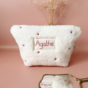 La conception de la trousse permet aux produits de bébé de tenir debout à l'intérieur, ce qui facilite l'organisation et l'accès aux produits essentiels. Liniment, couches, lait de toilette, cotons, autant de produits pour un si petit bout de chou, que vous aurez besoin d'une grande trousse de toilette spacieuse. Fabriquée en fausse fourrure sherpa écrue, elle est joliment brodée avec de petits coeurs rose. Cette trousse est à la fois douce et durable. L'intérieur de la trousse est doublé avec un tissu imperméable. Ls liquides renversés ne pénètrent pas à l'extérieur, protégeant ainsi les autres articles dans le sac à langer. Personnaliser votre trousse de toilette, au doux prénom de bébé ! La personnalisation est réalisée à la main sur le devant de la trousse de toilette avec un fil du coloris de votre choix.  C'est un plaston qui agrémente le devant de la trousse avec le prénom de bébé. Fièrement fabriquée en France, cette trousse de toilette est le fruit d'un savoir-faire artisanal et d'une attention particulière aux détails. Zoom sur le petit curseur de la fermeture à glissière ! Découvrez notre gamme entière de produits autour du bain : de belles et grandes capes de bain XXL, langes et débarbouillettes. 