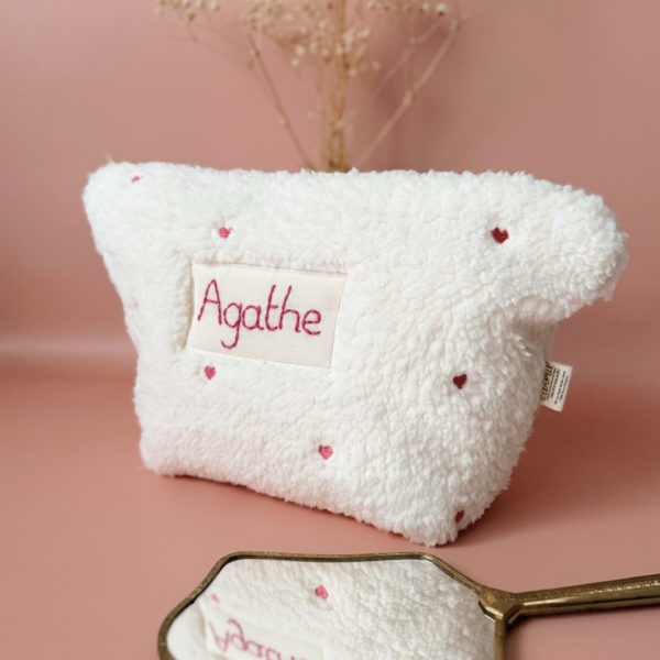 La conception de la trousse permet aux produits de bébé de tenir debout à l'intérieur, ce qui facilite l'organisation et l'accès aux produits essentiels. Liniment, couches, lait de toilette, cotons, autant de produits pour un si petit bout de chou, que vous aurez besoin d'une grande trousse de toilette spacieuse. Fabriquée en fausse fourrure sherpa écrue, elle est joliment brodée avec de petits coeurs rose. Cette trousse est à la fois douce et durable. L'intérieur de la trousse est doublé avec un tissu imperméable. Ls liquides renversés ne pénètrent pas à l'extérieur, protégeant ainsi les autres articles dans le sac à langer. Personnaliser votre trousse de toilette, au doux prénom de bébé ! La personnalisation est réalisée à la main sur le devant de la trousse de toilette avec un fil du coloris de votre choix.  C'est un plaston qui agrémente le devant de la trousse avec le prénom de bébé. Fièrement fabriquée en France, cette trousse de toilette est le fruit d'un savoir-faire artisanal et d'une attention particulière aux détails. Zoom sur le petit curseur de la fermeture à glissière ! Découvrez notre gamme entière de produits autour du bain : de belles et grandes capes de bain XXL, langes et débarbouillettes. 