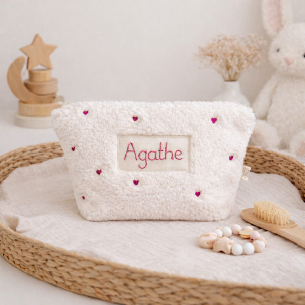 La conception de la trousse permet aux produits de bébé de tenir debout à l'intérieur, ce qui facilite l'organisation et l'accès aux produits essentiels. Liniment, couches, lait de toilette, cotons, autant de produits pour un si petit bout de chou, que vous aurez besoin d'une grande trousse de toilette spacieuse. Fabriquée en fausse fourrure sherpa écrue, elle est joliment brodée avec de petits coeurs rose. Cette trousse est à la fois douce et durable. L'intérieur de la trousse est doublé avec un tissu imperméable. Ls liquides renversés ne pénètrent pas à l'extérieur, protégeant ainsi les autres articles dans le sac à langer. Personnaliser votre trousse de toilette, au doux prénom de bébé ! La personnalisation est réalisée à la main sur le devant de la trousse de toilette avec un fil du coloris de votre choix.  C'est un plaston qui agrémente le devant de la trousse avec le prénom de bébé. Fièrement fabriquée en France, cette trousse de toilette est le fruit d'un savoir-faire artisanal et d'une attention particulière aux détails. Zoom sur le petit curseur de la fermeture à glissière ! Découvrez notre gamme entière de produits autour du bain : de belles et grandes capes de bain XXL, langes et débarbouillettes. 