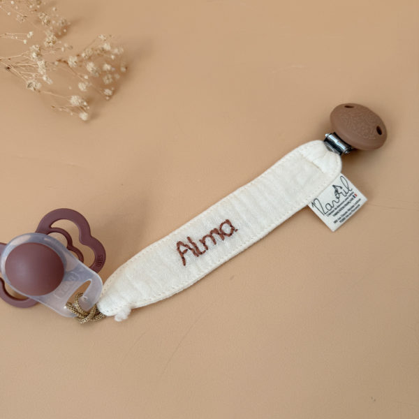 Élégante, douce et sécurisée, notre attache tétine en gaze de coton écrue accompagne bébé au quotidien tout en ajoutant une touche délicate à sa tenue. Chaque modèle est personnalisable avec le prénom de bébé, soigneusement brodé à la main, pour créer un accessoire unique et plein de sens. Dotée d’un lien doré raffiné pour fixer la tétine et d’une pince ronde en silicone embossé, elle allie esthétique et praticité.